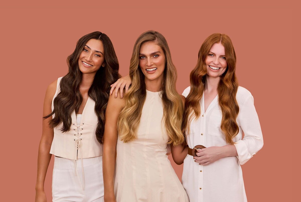 Three women with long, styled hair in brunette, blonde, and red shades, wearing neutral-toned outfits, posing against a terracotta background.