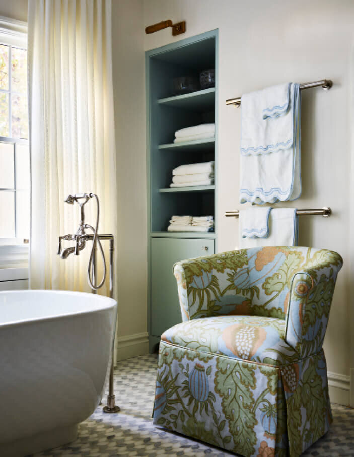 Interior design in Boston features a bathroom with Christopher Farr Cloth upholstered slipper chairs, a freestanding tub, blue cabinets, and brass lighting.