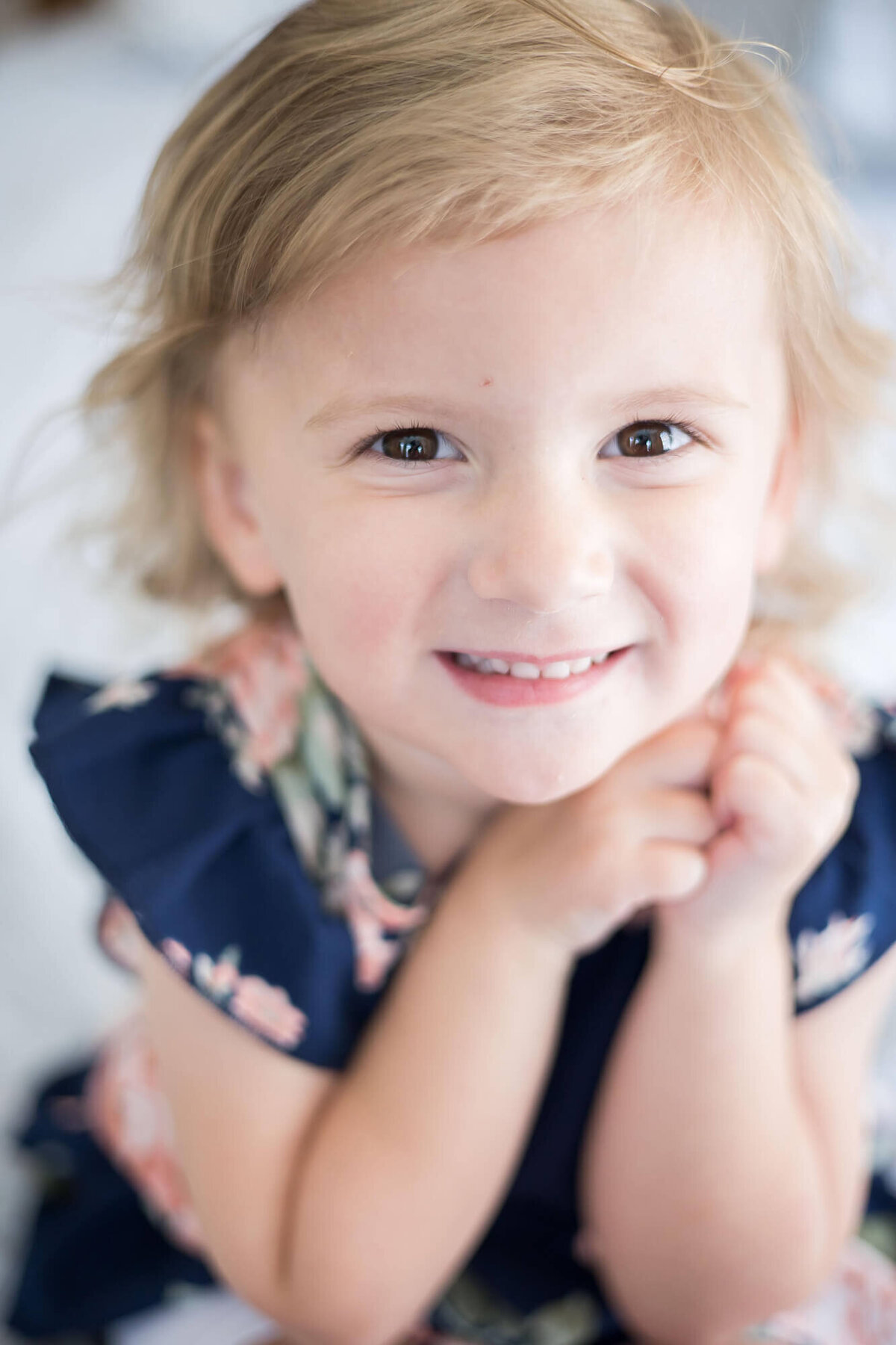 Cute blonde three year old girl in a blue dress smiling with her fists by her chin