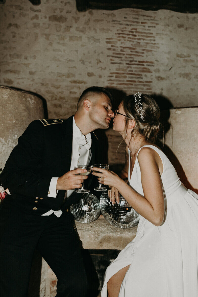 Deux mariés, l'homme en uniforme noir trinquant avec des coupes de champagnes, appuyés sur des boules à facette, capturés par Laura, lors d'un shooting photo mariage en vendée.