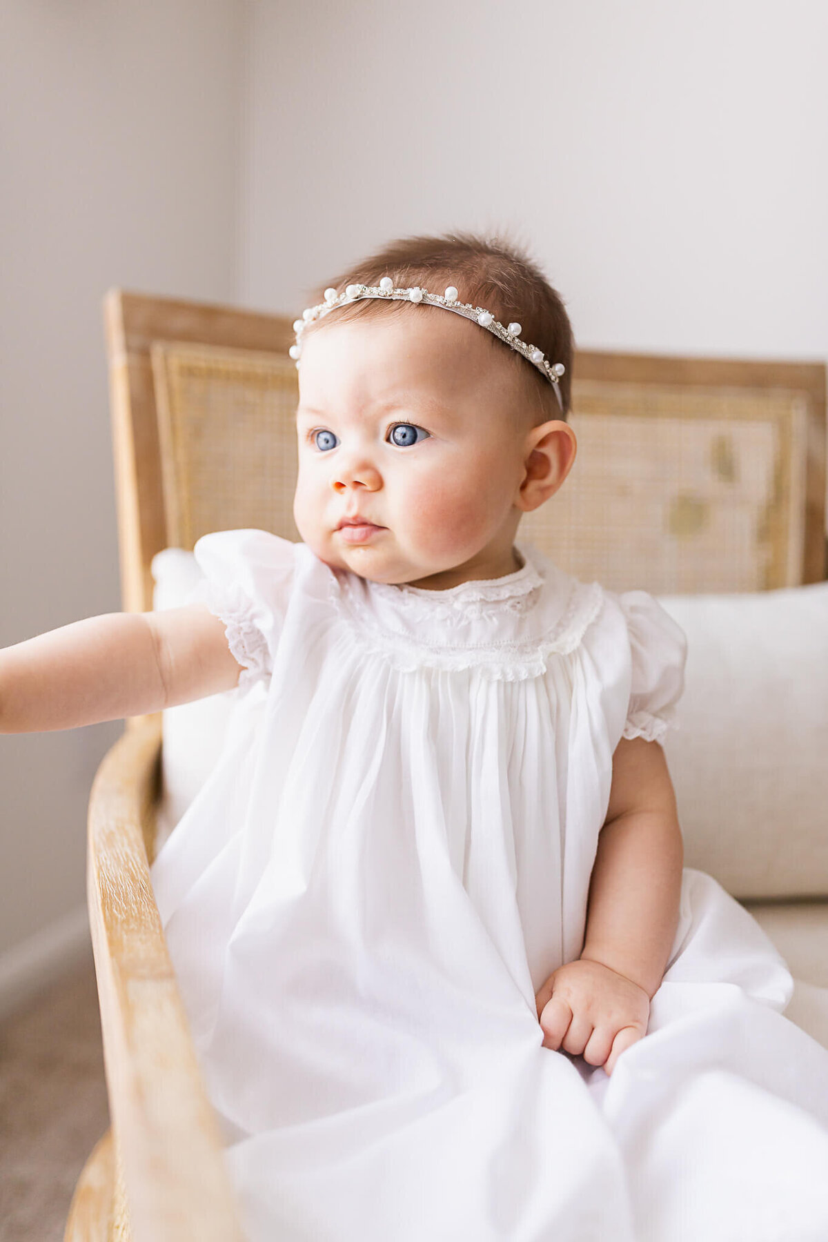 Six month old baby sitting in a chair in a white dedication dress and pearls