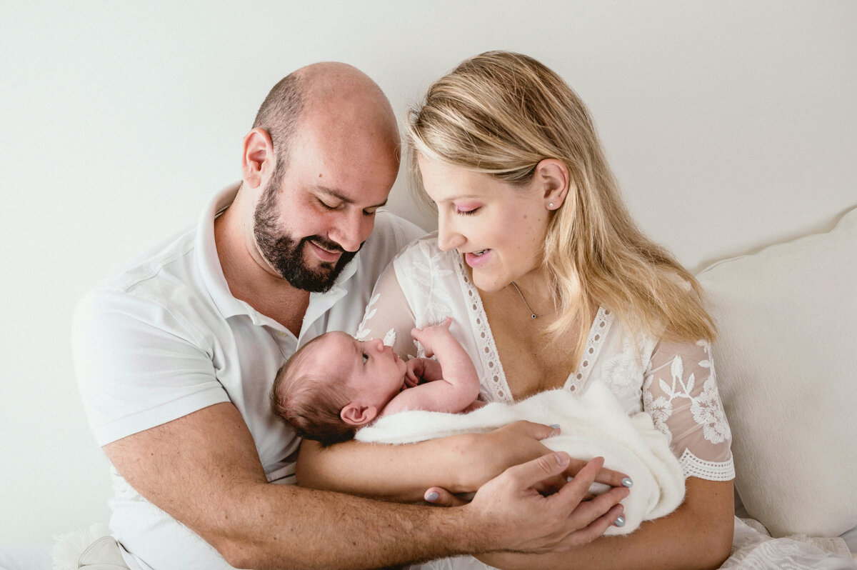 A blond mom and balding father holding their newborn baby  on a bed and looking down lovingly.