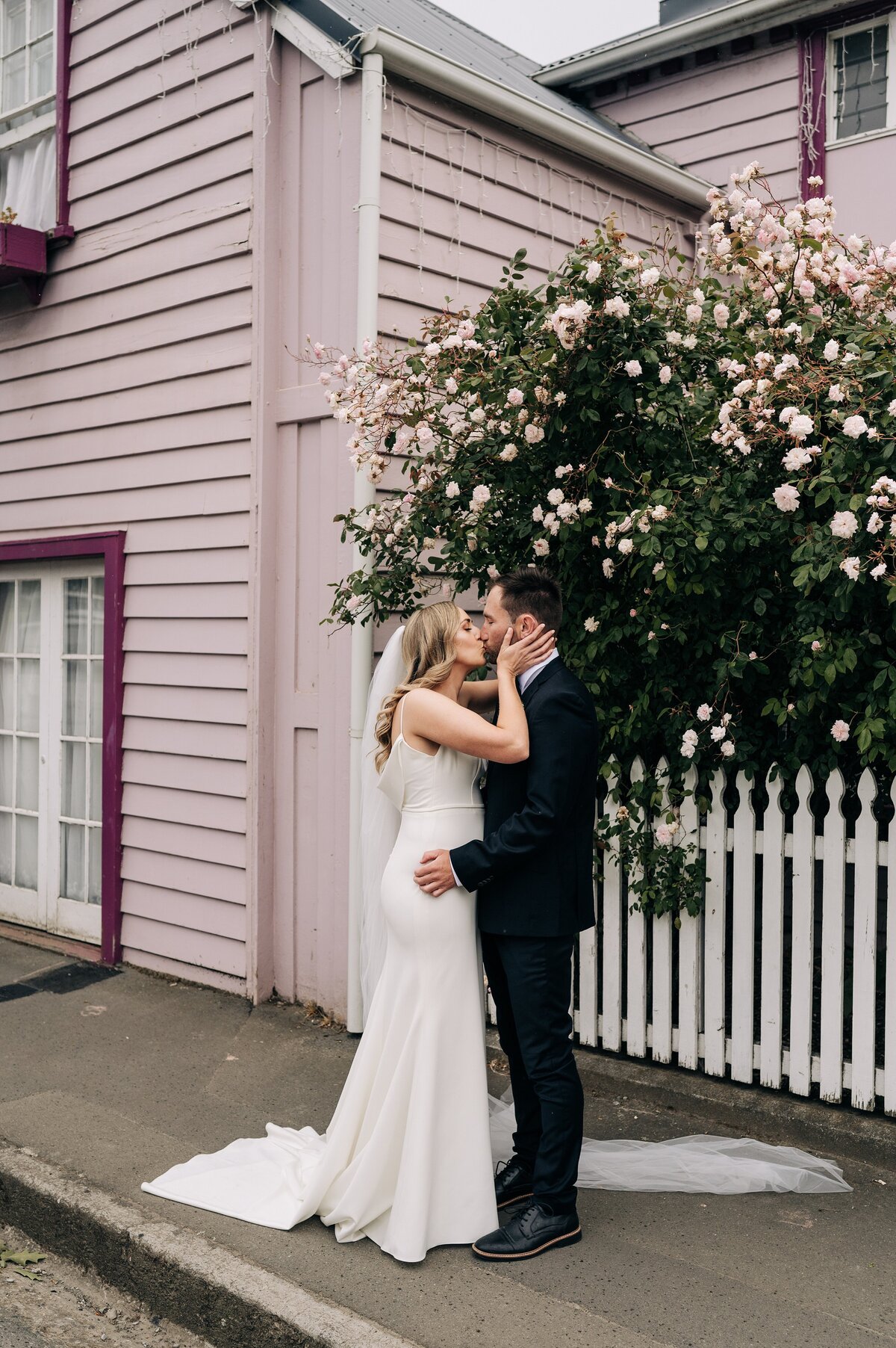 bride and groom kiss in front of pink house with pink floral vine in akaroa new zealand on their wedding day