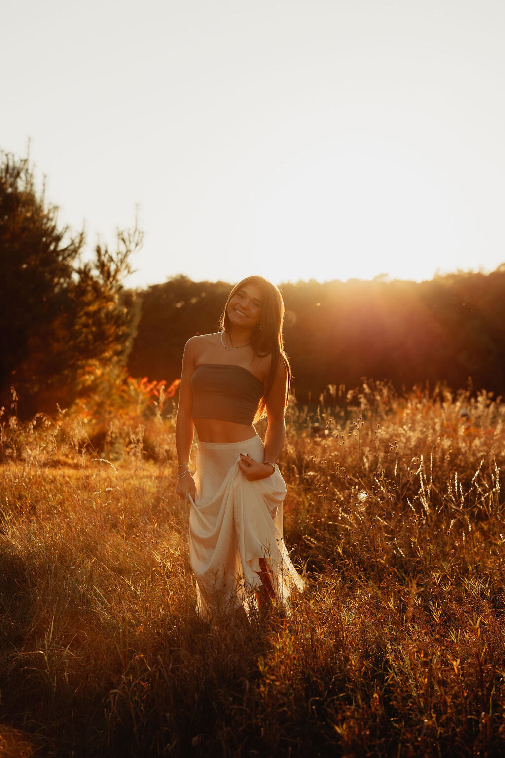 A woman stands in a sunlit field at sunset, holding the hem of her long skirt. She wears a strapless top and smiles softly. Captured by a New England brand photographer, the golden hour light creates a warm glow around her and the surrounding grass, with trees faintly visible in the background.