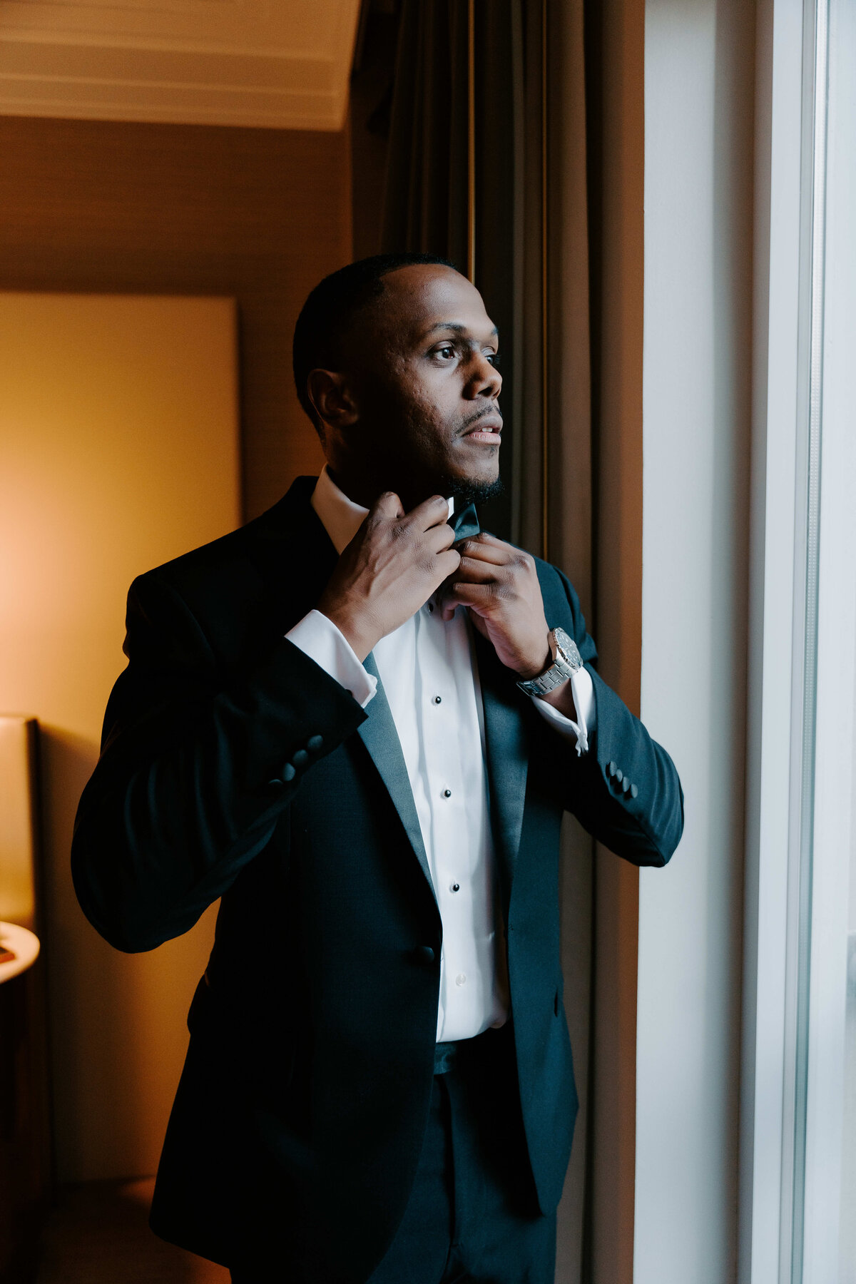 Groom getting ready on wedding day, adjusting his bow tie while looking out the window in a classic black tuxedo.