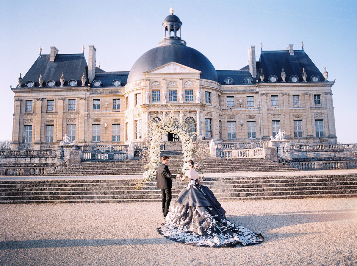 Captured in the romantic setting of Chateau Vaux le Vicomte in Paris, France, this breathtaking photo showcases one of the top destination wedding venues in the country. Expertly photographed by Audrey from East West Films Videographers, the image highlights a couple standing elegantly in front of an ornate, historic chateau adorned with a grand dome. The contrast between the woman's intricate black and white gown and the man's sharp black suit adds a touch of timeless sophistication to the scene. The couple is beautifully framed by a lush floral archway under the clear blue skies, creating a picturesque backdrop for a truly unforgettable moment.
