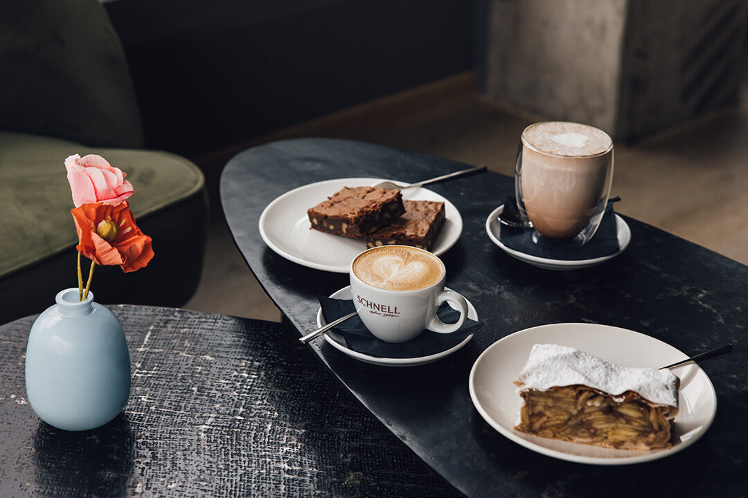 Zwei Schokobrownies, ein Stück Alt-Wiener Apfelstrudel, eine Tasse Cappuccino und ein Glas mit Latte Macchiato auf einem kleinen schwarzen Tisch beim SCHNELL in Lauterach