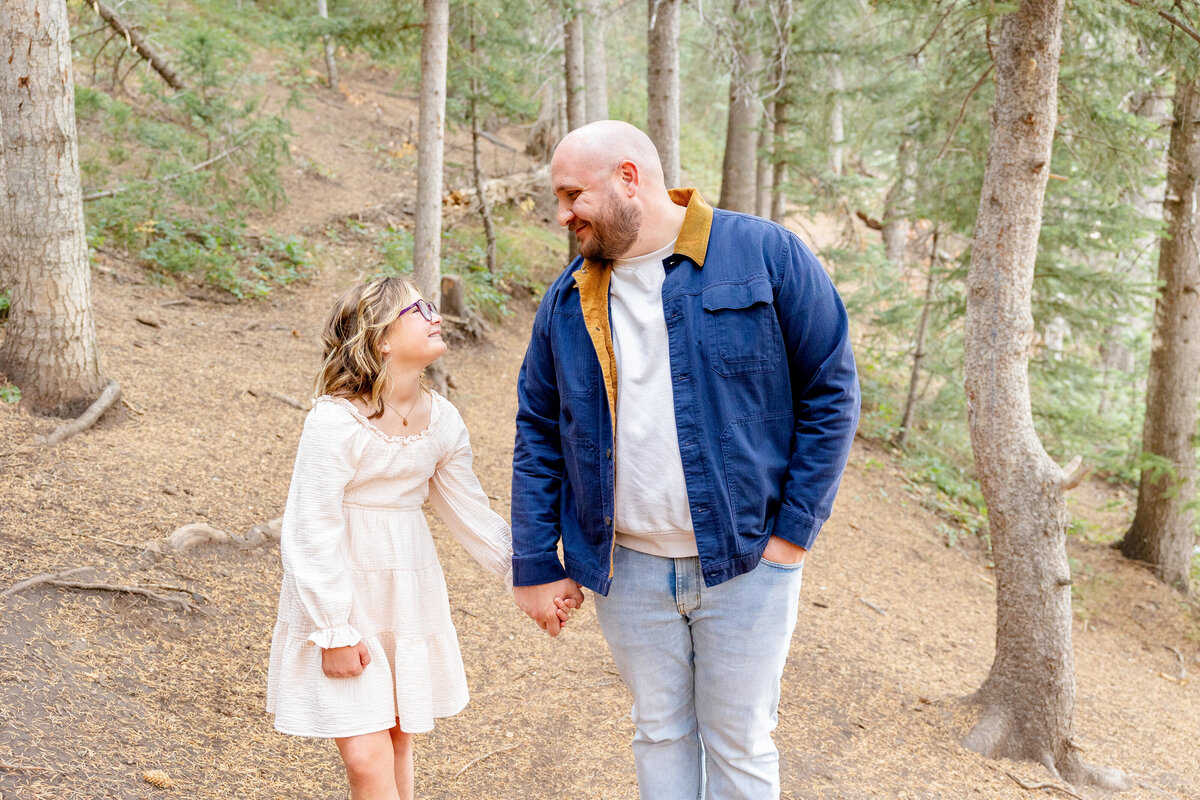 A father in a navy jacket and his daughter in a cream dress hold hands and share a warm smile as they walk together on a dirt path surrounded by trees.