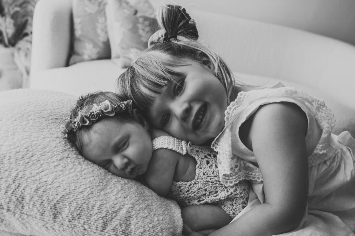 a sister that is hugging her newborn sister that is sleeping on a couch in a black and white photo