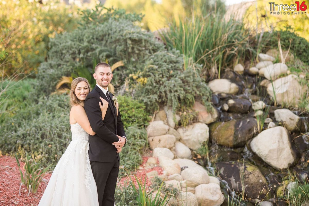 Bride embraces her Groom from behind as they pose for wedding photos next to a small waterfall rolling down large stones