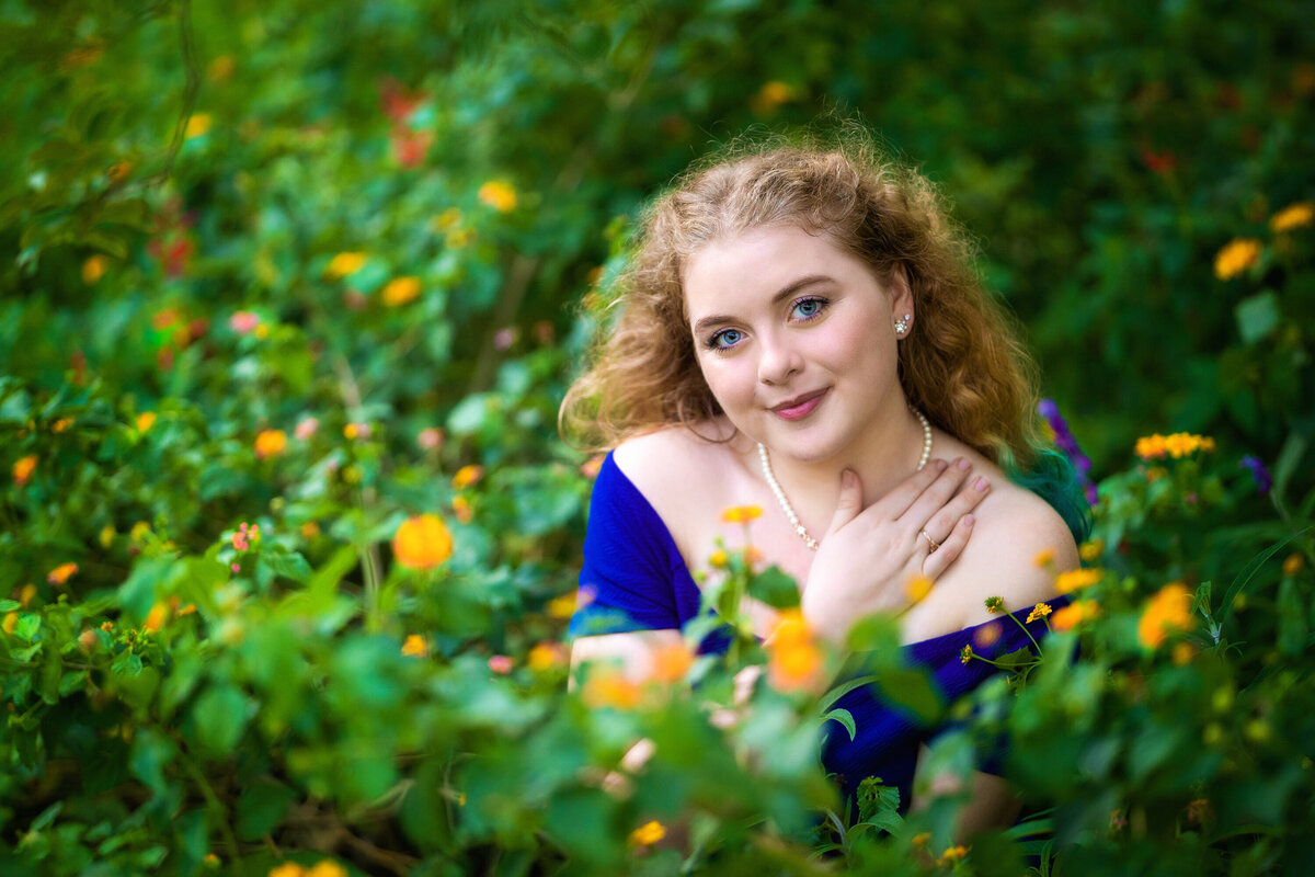 Senior girl with curly light brown hair in a patch of yellow flowers.  She is looking at the camera with a slight smile.  Her hand is raised towards her neck.  She is wearing a royal blue off the shoulder dress.
