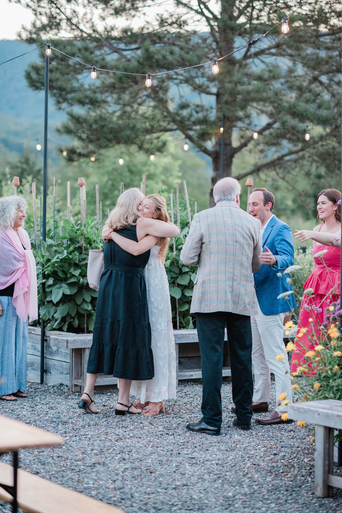 A bride in a white dress embraces a guest in a black dress during an outdoor welcome gathering, with string lights overhead and a green mountain landscape in the background.