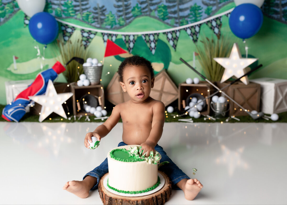 Golf themed cake smash in West Palm Beach and Jupiter, FL photography studio. Baby boy is sitting behind a green and white cake with a golf ball in one hand, the other hand playing with icing. He is looking at the camera. In the background there is a golf backdrop, clubs, and golf balls.