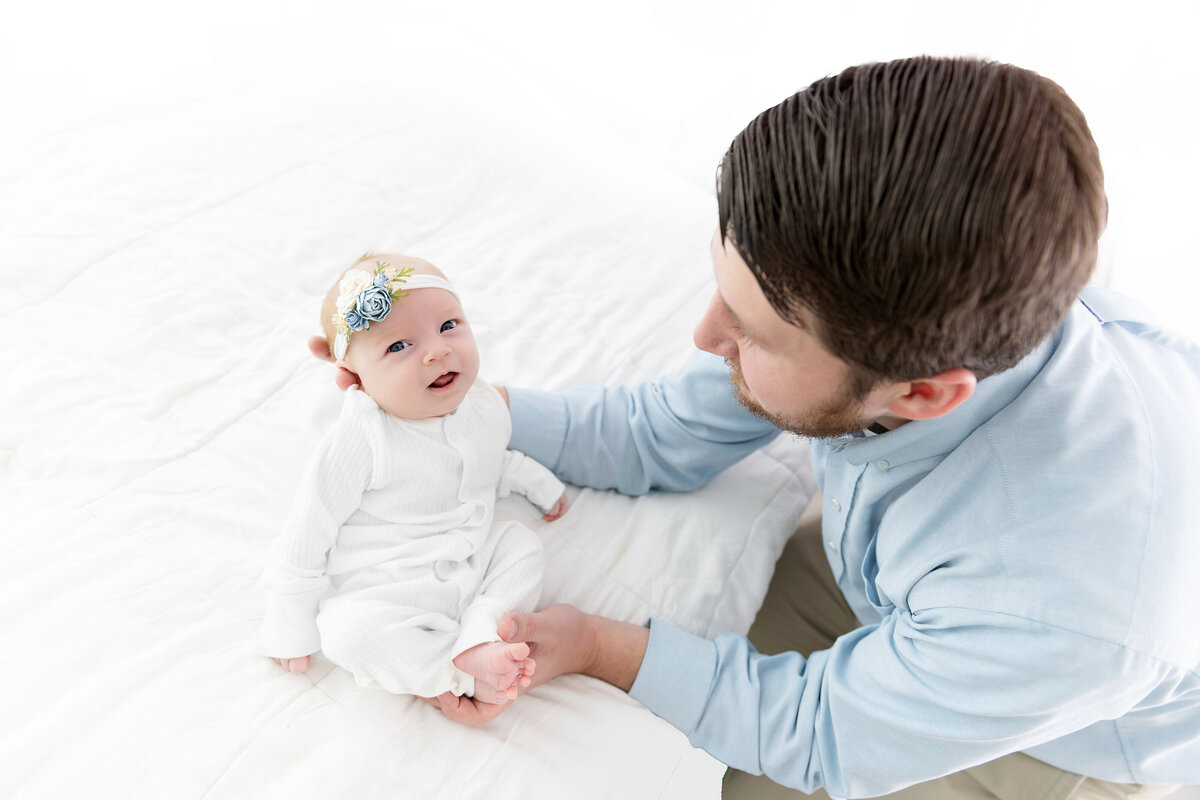 A father poses his newborn baby daughter on a bed in a white onesie as directed by an Atlanta newborn photographer