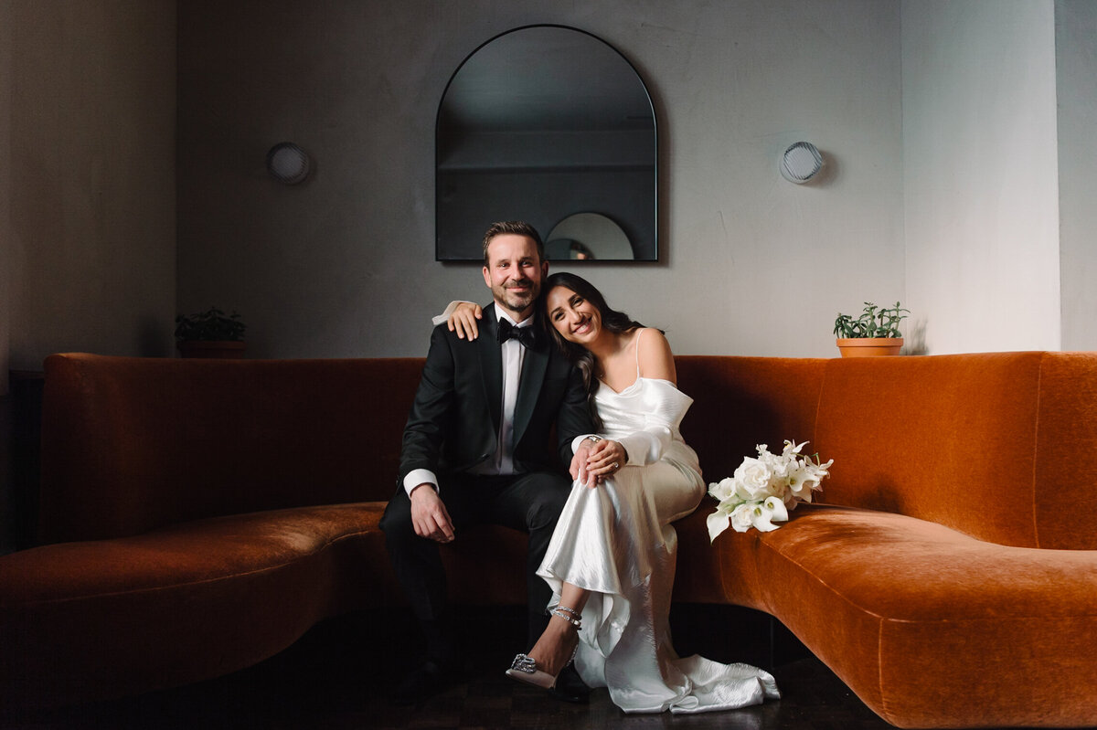A happy bride and groom sitting on an orange sofa at La Banane Restaurant in Toronto