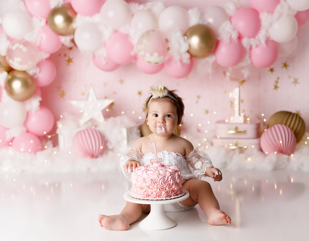 Pretty in Pink themed cake smash. Baby girl in cream lace onesie in front of a pink ombre rosette cake. look at the camera with cake on her face. In the background, there is a balloon arch of various shades of pink, suitcases, and faux clouds.
