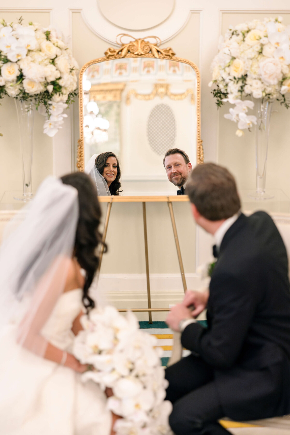 A bride and groom sit on the floor, smiling playfully at their reflection in a large, ornate mirror. Two large floral arrangements flank the mirror, creating an elegant backdrop. The bride holds a bouquet of white flowers.
