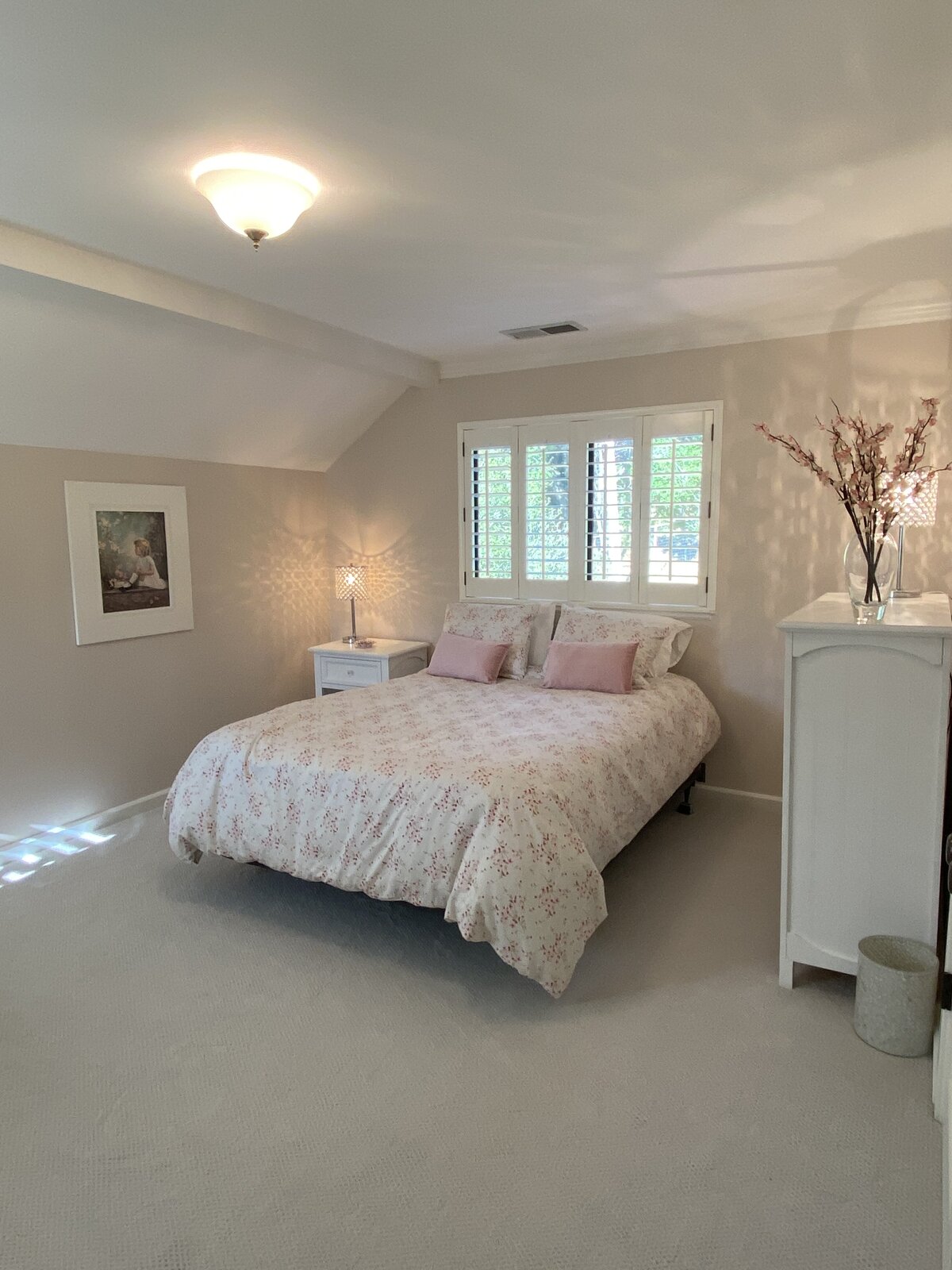A cozy, updated bedroom with neutral walls, a sloped ceiling, plantation shutters, and soft pink bedding after a home improvement project.
