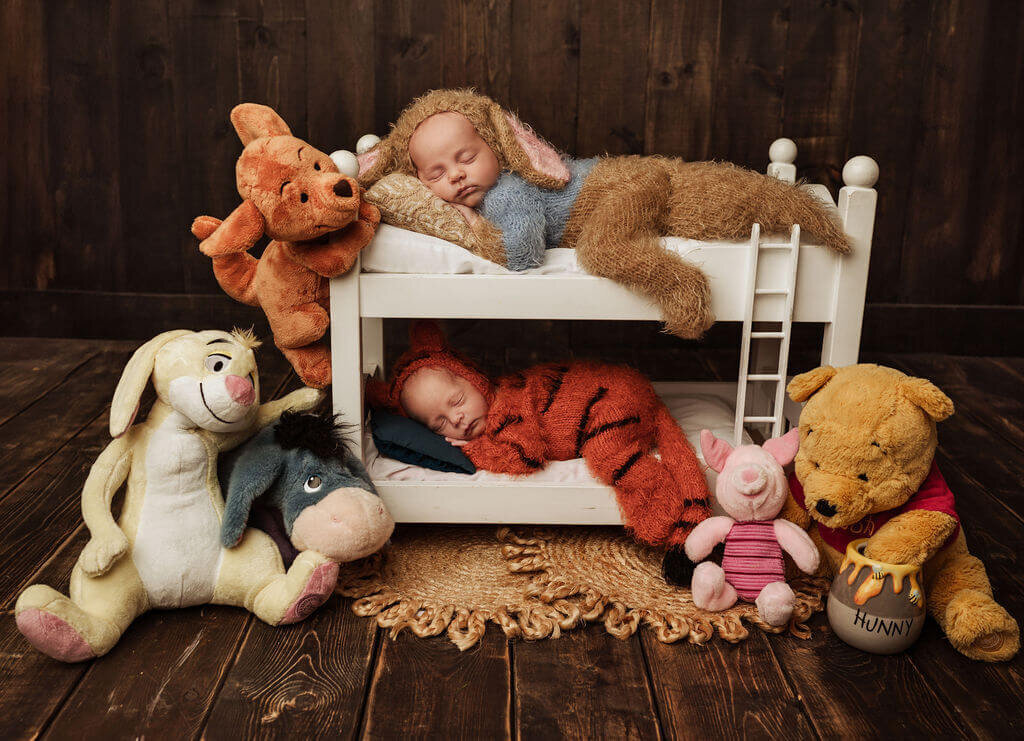 Twin newborn babies at their Toronto Newborn photo shoot dressed in soft knit onezies as Roo and Tigger from the beloved Winnie The Pooh cartoon, laying on a bunk bed surrounded by plush stuffies from the show.