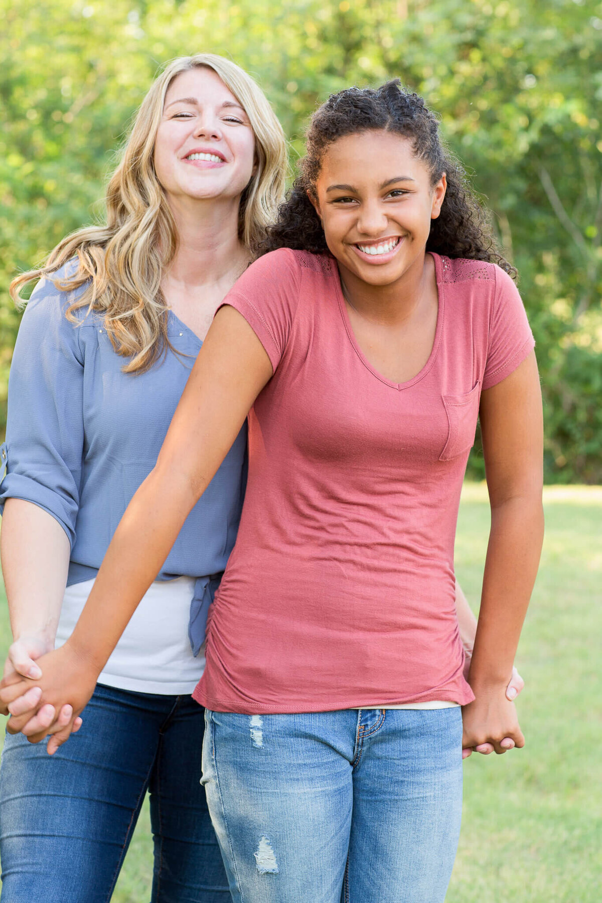 A woman and a girl smile brightly, holding hands amidst a lush green backdrop. The woman dons a blue top and jeans, while the girl sports a pink shirt and jeans. Captured by a Fayetteville NC family photographer, this moment radiates joy and connection.