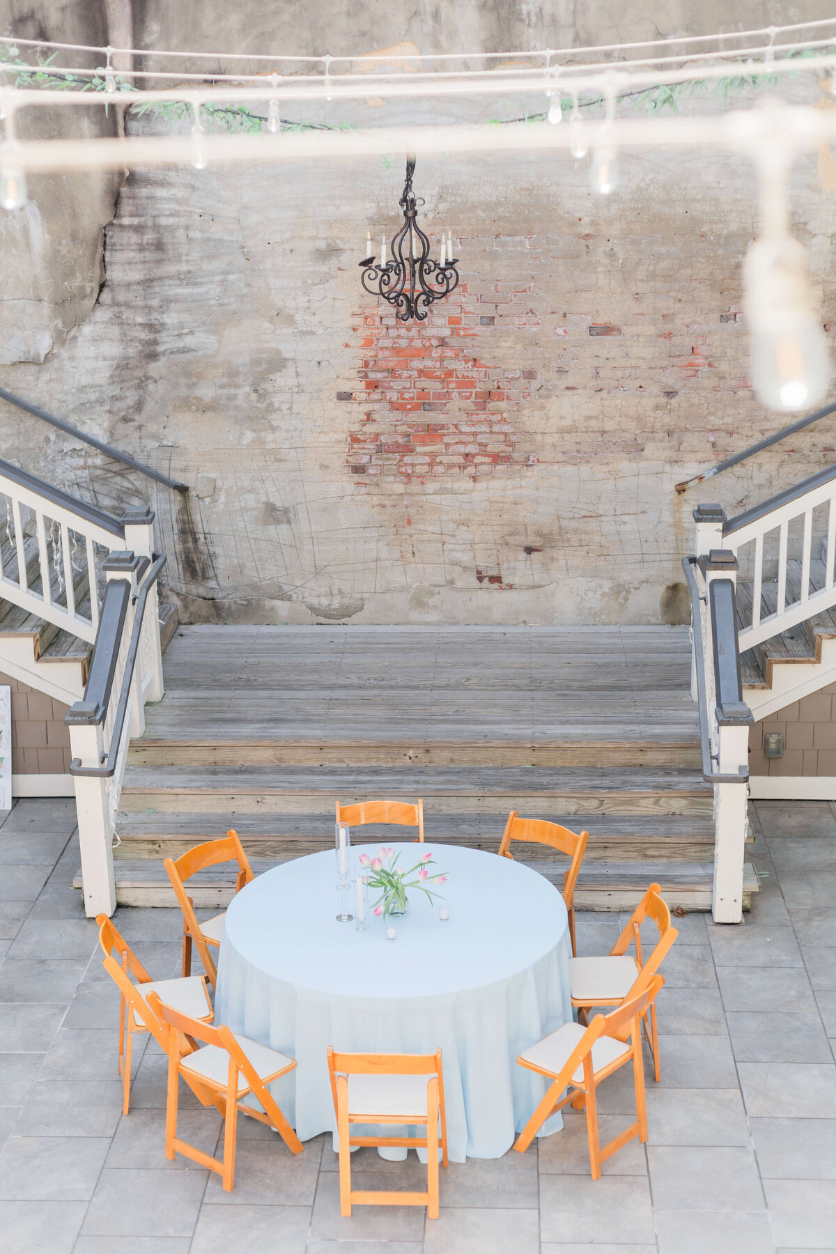 A round table with a light blue tablecloth and six orange wooden chairs sits on a patio in front of worn stone steps and a brick wall with a black chandelier hanging above. A centerpiece with flowers is on the table.