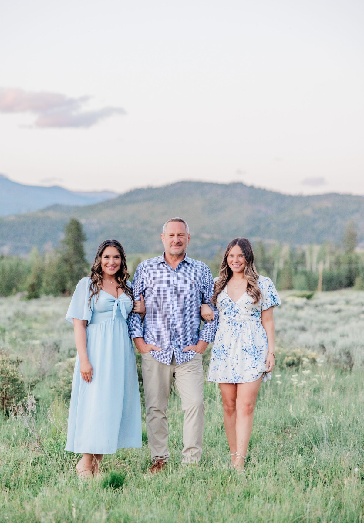 A dad in a blue shirt stands with his wife and daughter hanging on his arms while walking a mountain meadow