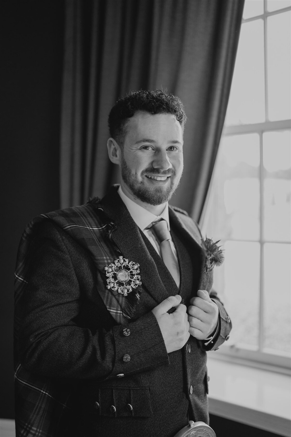 A groom stands by a large old window and smiles candidly as he gets his photograph taken. The courtyard of Logie Country House is visible through the window. The groom wears traditional Scottish attire.