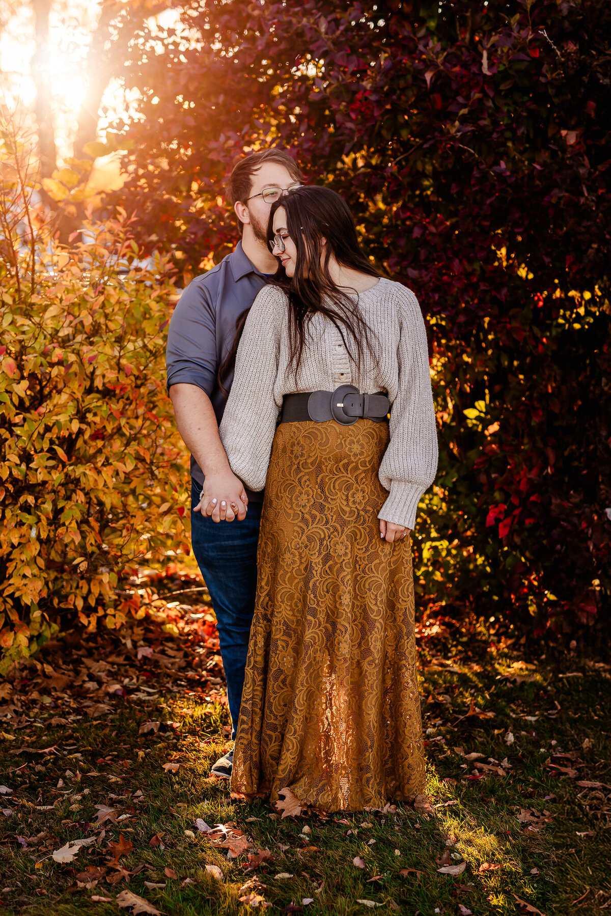 Girl in long green dress and tan sweater looking back at her fiancee in fall foliage.