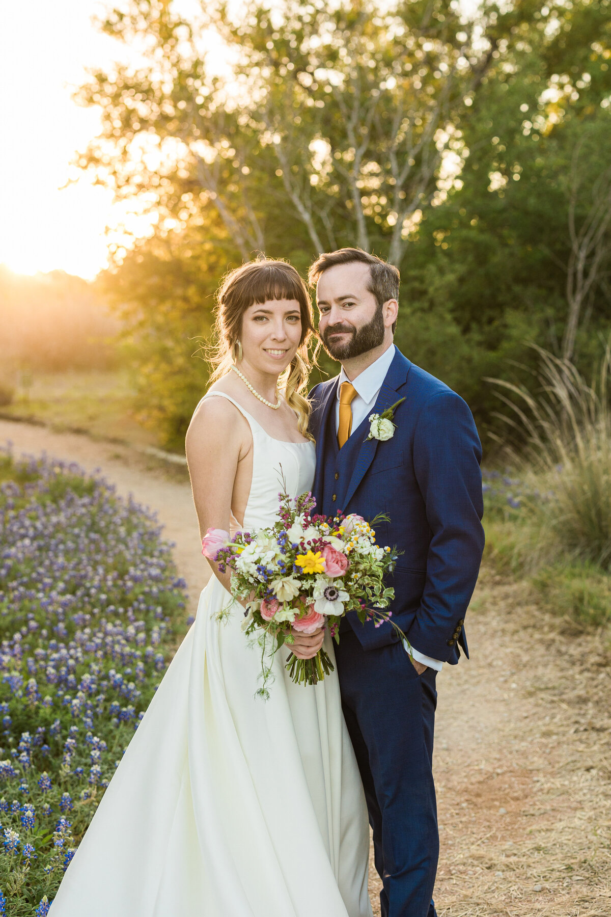 Portrait of a bride and groom at golden hour after their wedding ceremony at the Lady Bird Johnson Wildflower Center in Austin, Texas. The couples stands on a nature trail with many trees on one side and many bluebonnets on the other. The bride is on the left and is wearing a sleeveless, elegant, white dress and is holding a bouquet. The groom is on the right and is wearing a navy suit with an orange tie and boutonniere.