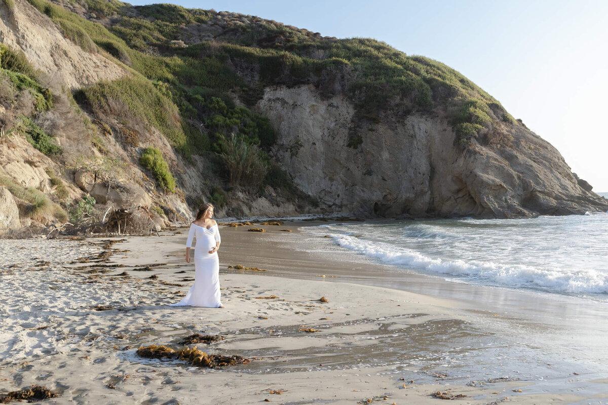A woman in a white dress stands on a sandy beach near the shoreline, gazing at the ocean. The backdrop features rocky cliffs covered with greenery. The scene is calm and sunny.
