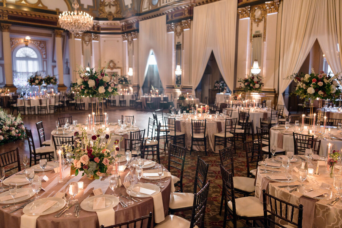 A luxurious banquet hall with round tables elegantly set for an event. Each table features floral centerpieces, candles, and neatly arranged dining ware. Large chandeliers hang from the ornate ceiling, and soft lighting creates a warm ambiance.