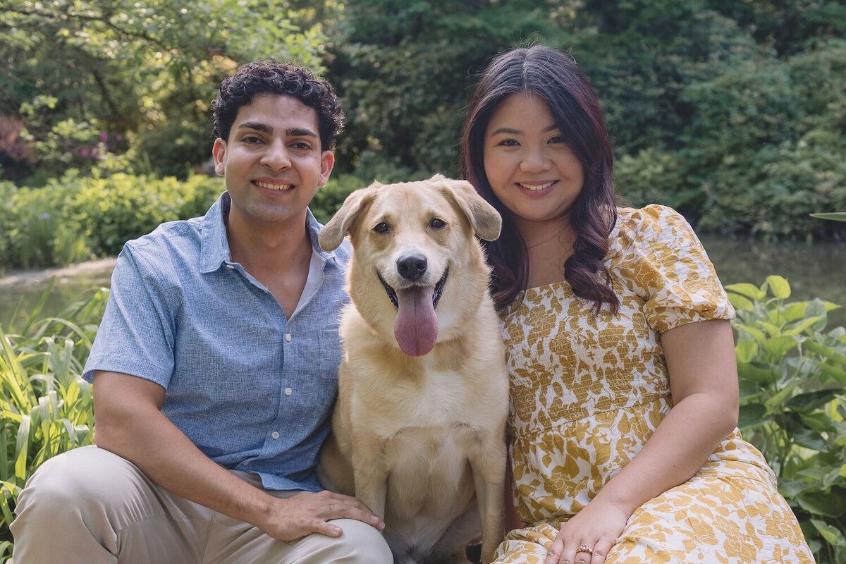 Couple smiling with their dog in a sunlit garden setting.