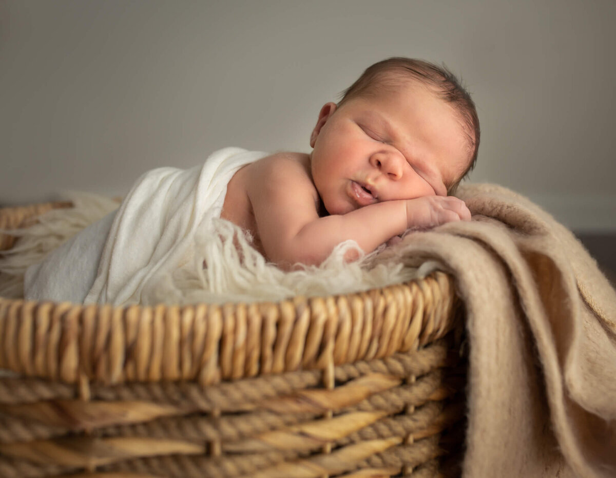 A newborn baby boy lays in a basket and snoozes with his hands under his cheeks