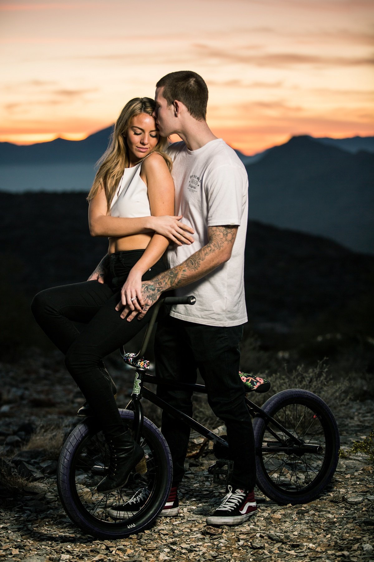 Bride to be sits on her fiance's bike handlebar as he whispers in her ear during an evening photo session