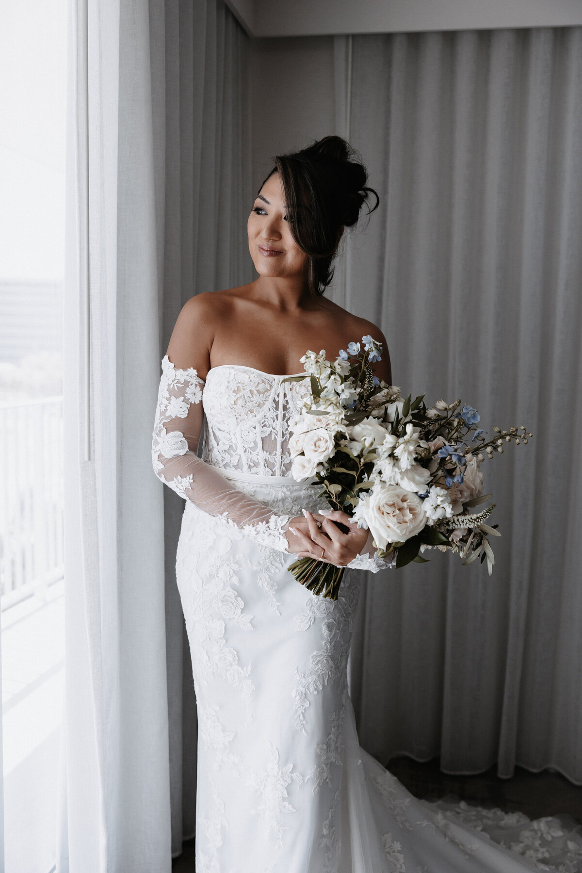 Bride holding flowers looking out the window in lace vera wang strapless dress with detached sleeves