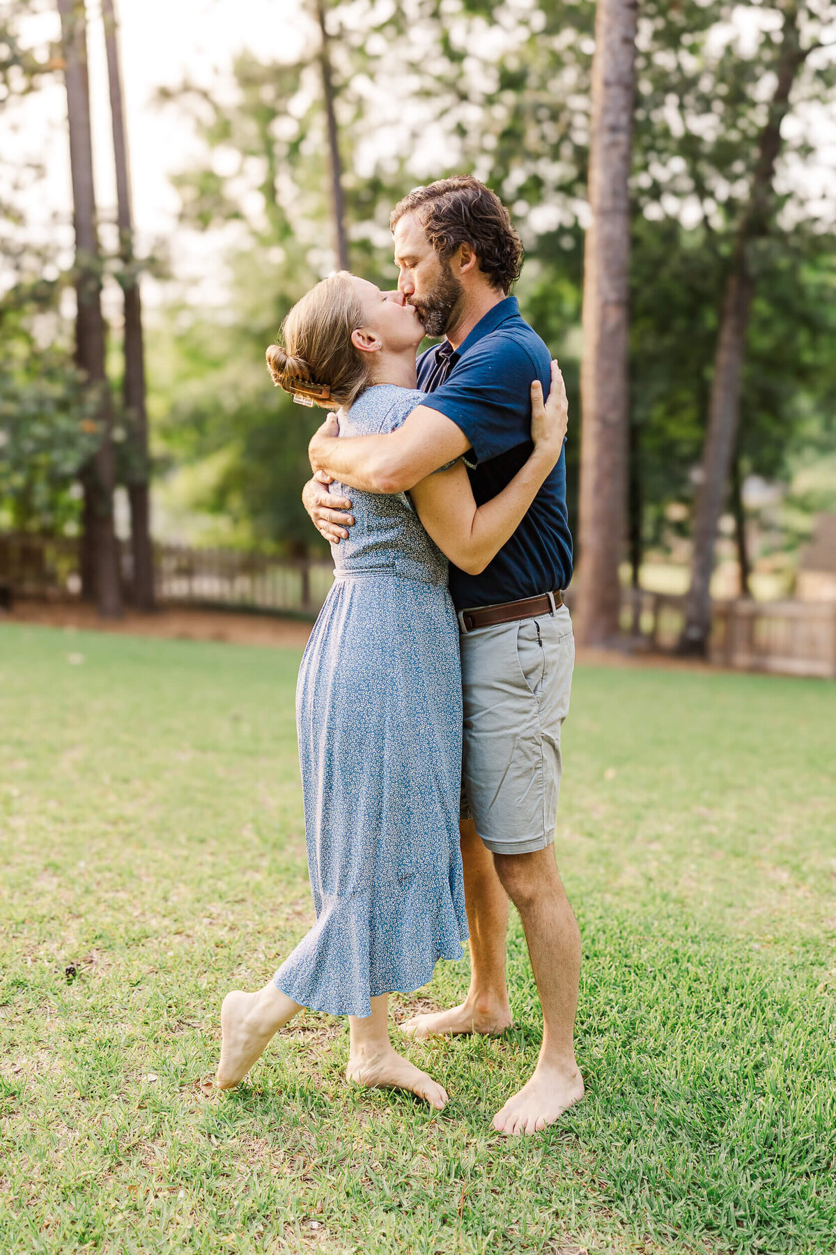 A man and woman are embracing and kissing in a grassy yard, captured by a Fayetteville NC family photographer. The woman is wearing a blue dress, and the man is in a navy shirt and khaki shorts. Both are barefoot, with trees visible in the background.