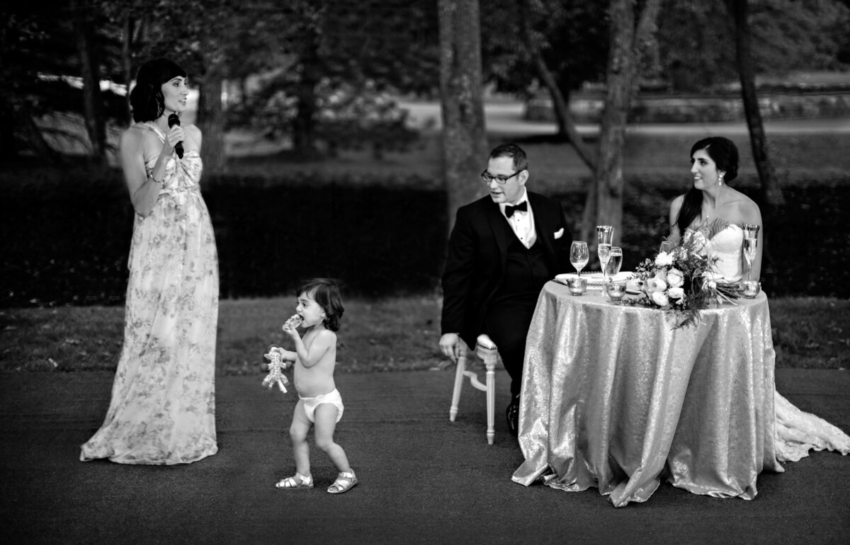 The matron of honor gives her speech as her daughter runs by after taking her dress off. The groom and bride are taken by surprise.