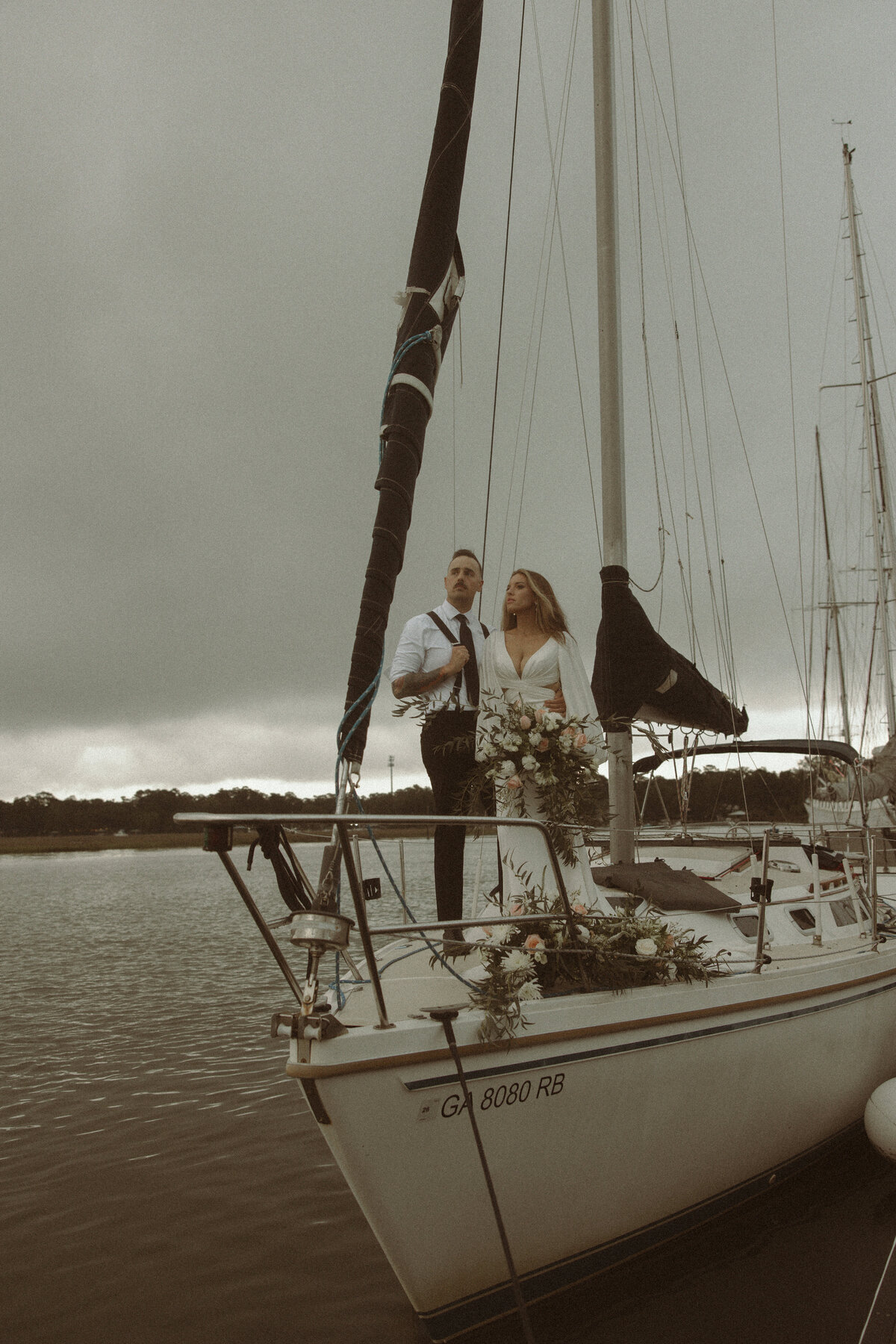 A couple stands on the bow of a sailboat adorned with fresh flowers, set against a moody overcast sky over calm waters.