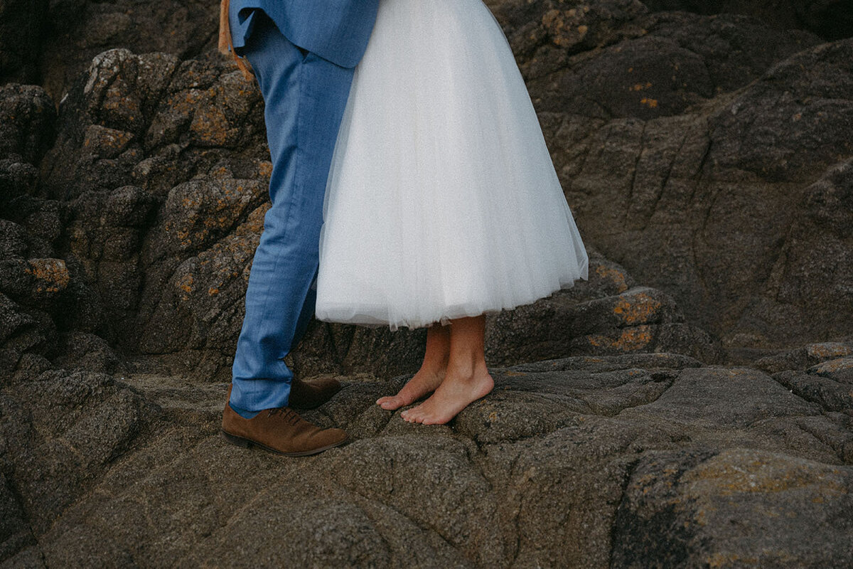 Plan rapproché sur les pieds nus des mariés, debout face à face sur les rochers de bord de mer. Scène saisie par Kamescop, photographe mariage vintage.