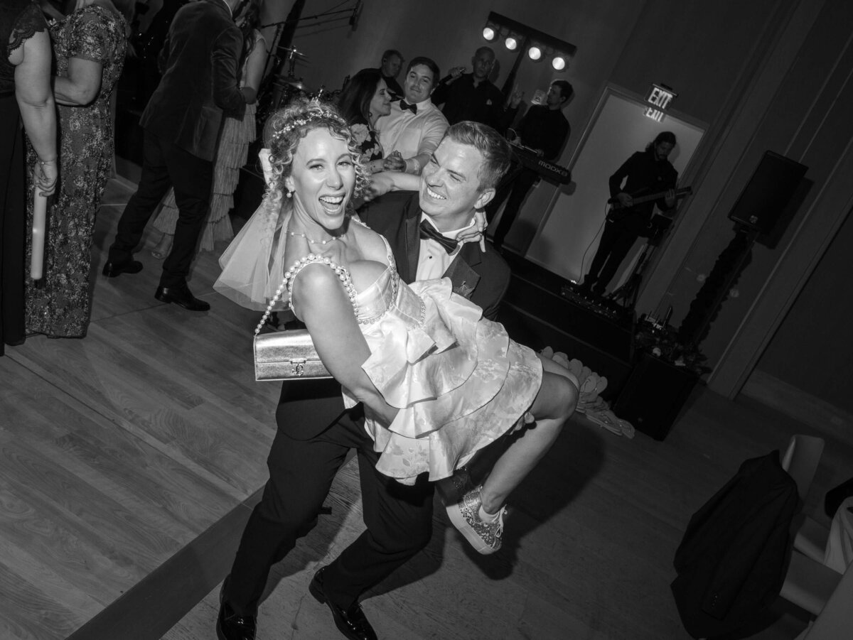 Groom carrying bride on the dance floor during wedding reception, both laughing and celebrating with guests.