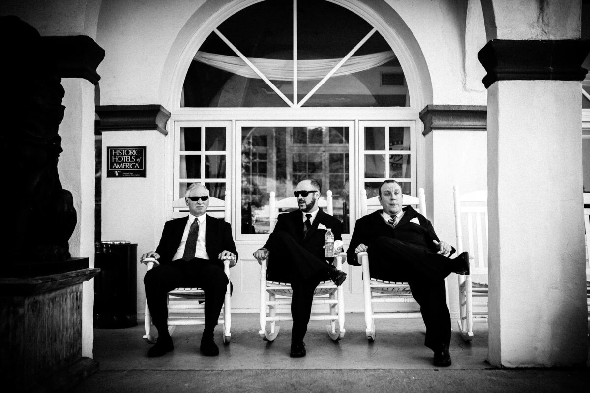 Three groomsmen sit nonchalantly in sunglasses on rocking chairs outside a classic venue