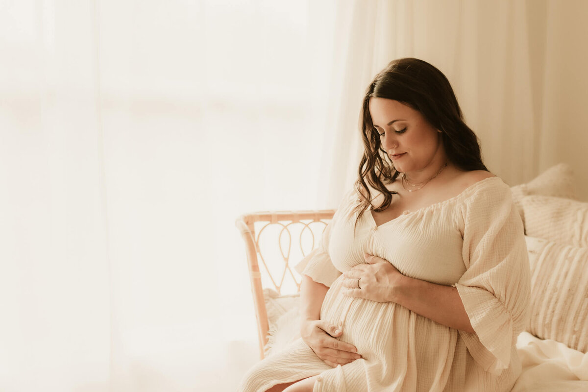 Mother holding her baby bump while sitting down near a window.
