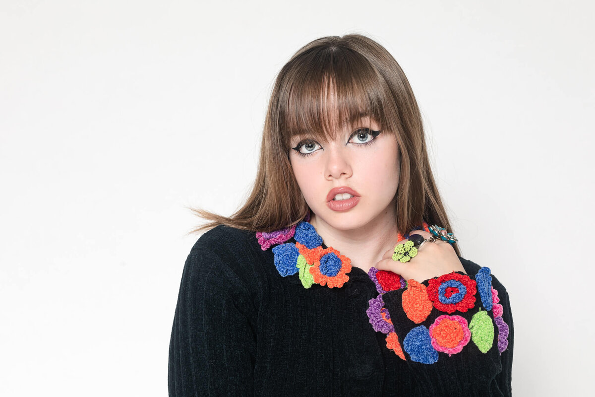 A close-up of a young brunette woman with bangs posing with her left hand touching her left shoulder, wearing a black sweater with colorful flowers on the sleeves, looking serious with her lips not touching as she's looking at the camera in a white studio.
