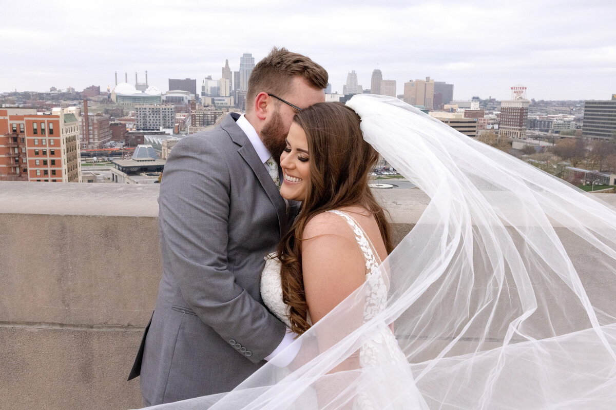 bride and groom embracing while standing on top of a tall building with city in the background