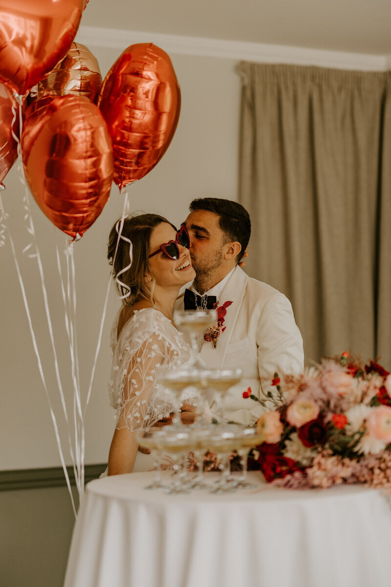 Mariée avec des lunettes en forme de coeur recevant un bisou sur la joue du marié. Structure de coupes de champagne, bouquet et ballons métalliques rouges au premier plan. Posent pour le workshop photographie de mariage.