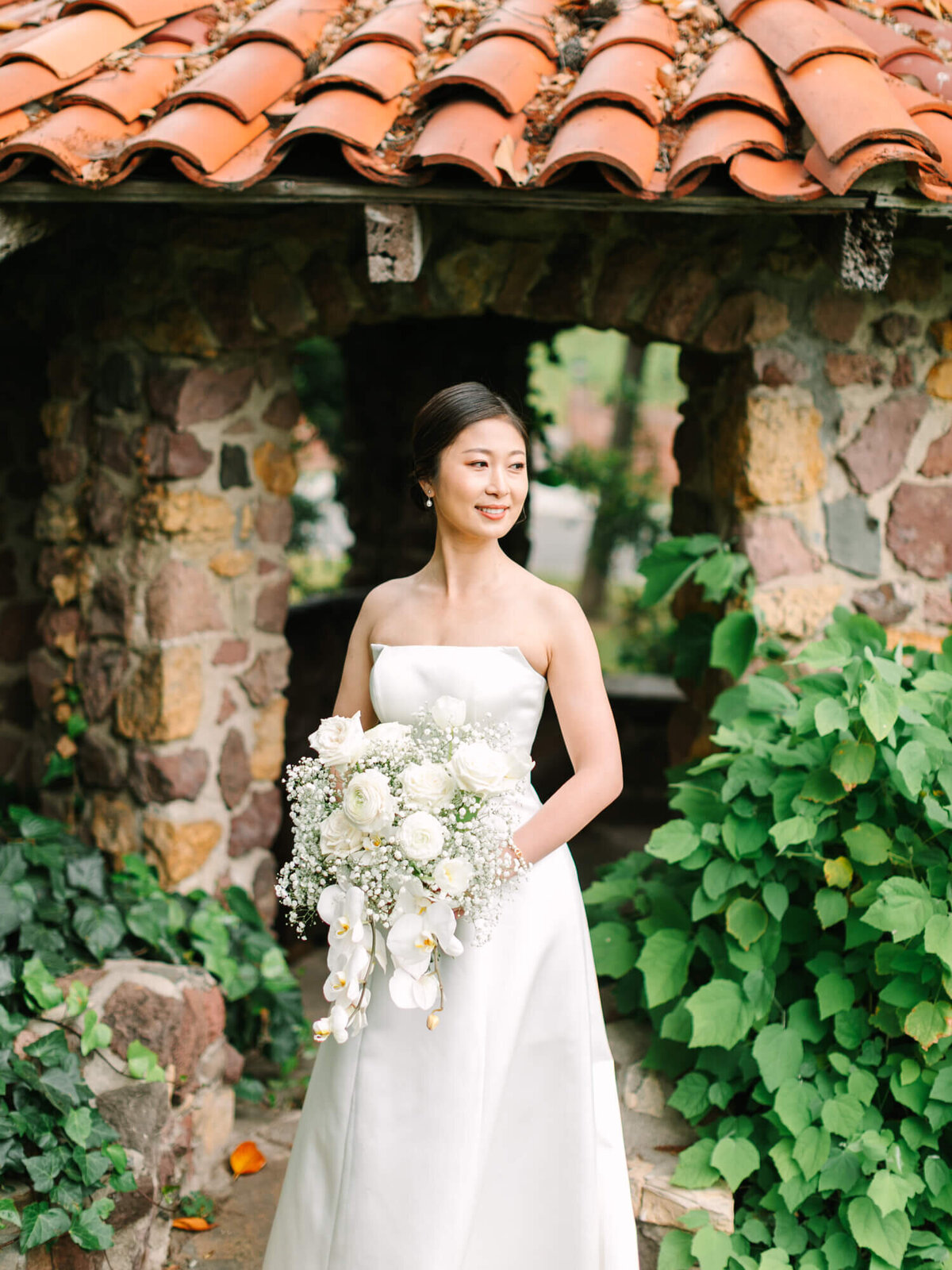 Bride in a strapless white gown holds a bouquet of white flowers, standing by a stone archway with greenery. The scene is serene and romantic.