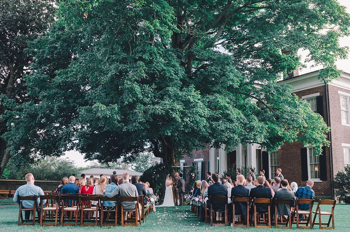 Brown fruit wood garden chairs set up underneath a large tree with wedding guests. The bride and groom exchange vows underneath a purple floral arbor. Rippavilla Plantation sits in the distance.