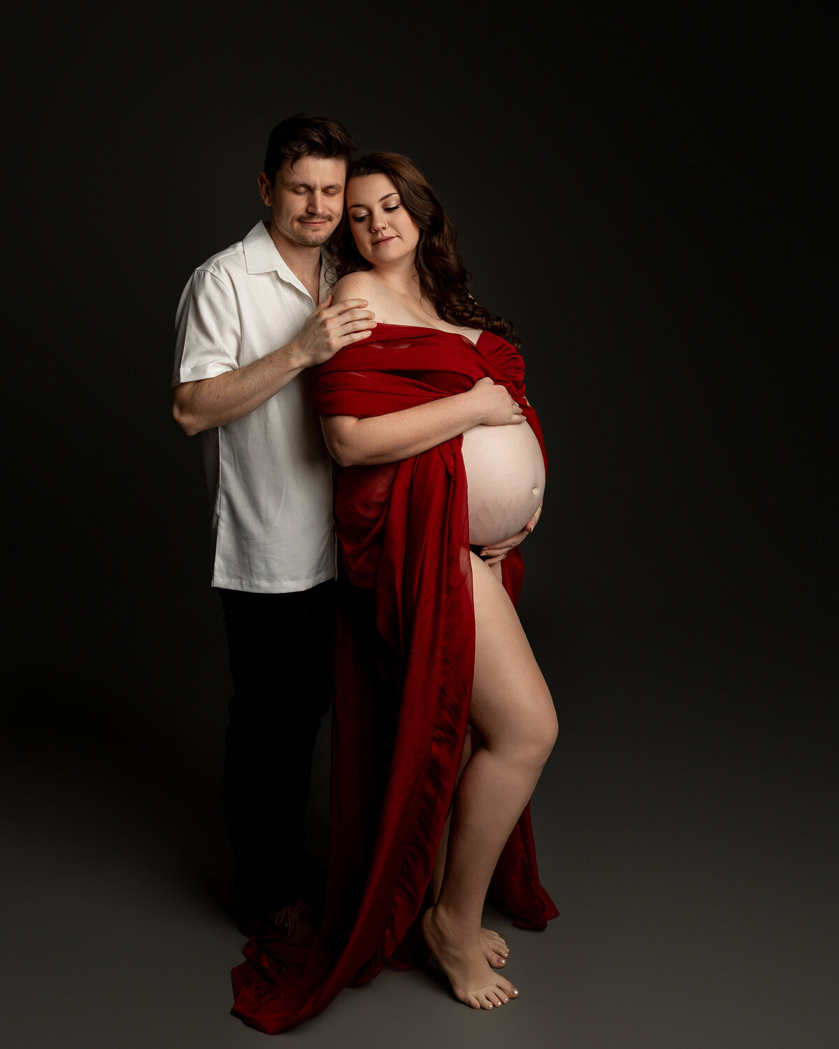 In a red dress, the expectant mother stands with her husband, posing side by side against a dark backdrop, capturing the beauty of their growing family. Captured by Inland Empire maternity photographer Kaitlyn Dawn Photography.