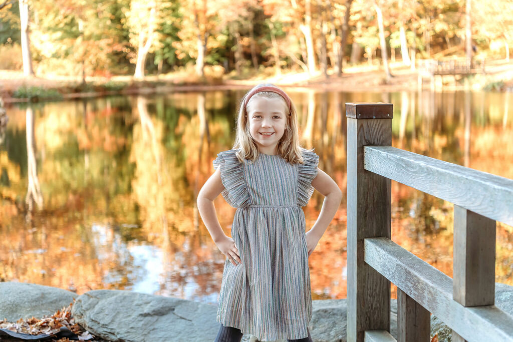Young girl in striped dress standing confidently by wooden fence during Connecticut family photographer lakeside autumn session