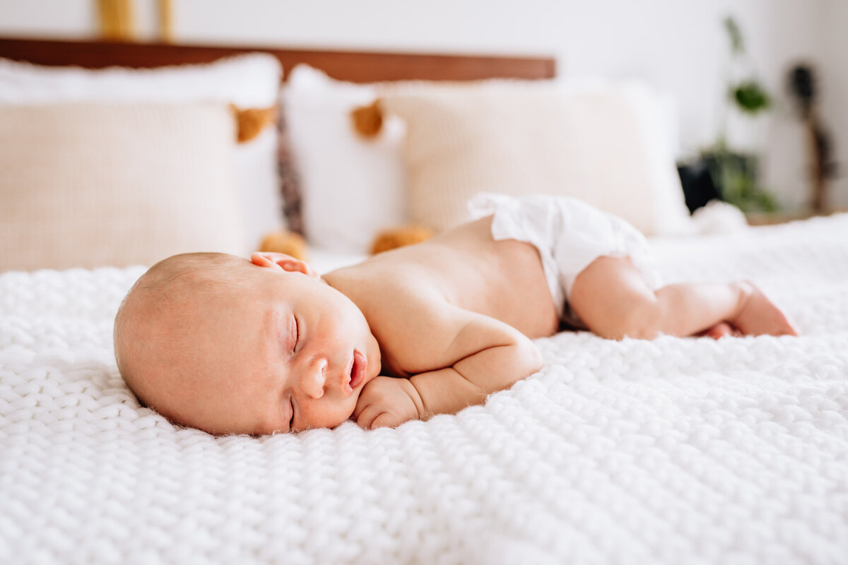A close-up photograph of a newborn baby peacefully sleeping on a white, textured blanket, wearing only a diaper. The background shows soft pillows and a bed.